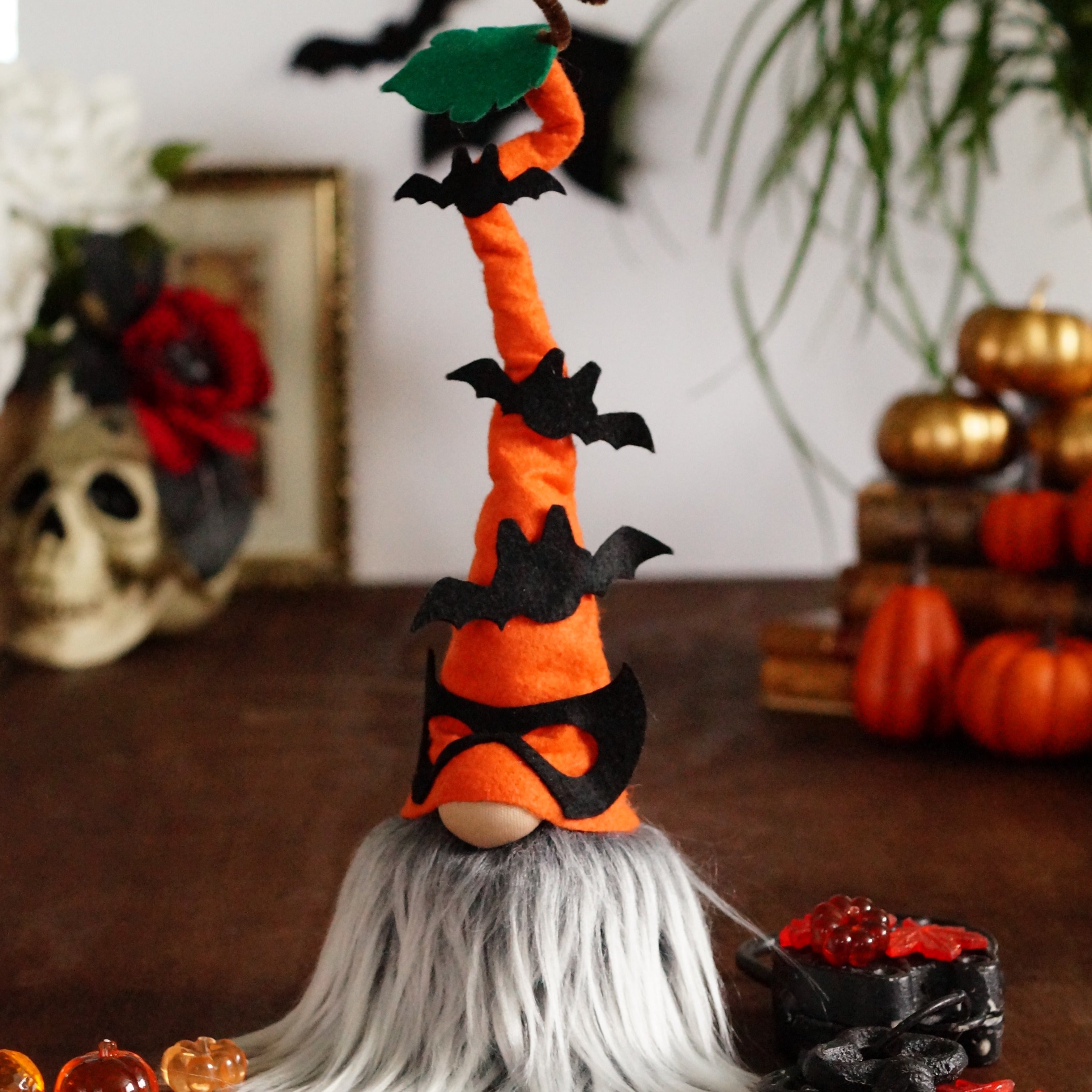 A decorative Halloween gnome, dressed in an orange outfit and wearing an orange hat adorned with bats, against a white background with books, pumpkins and a skull.