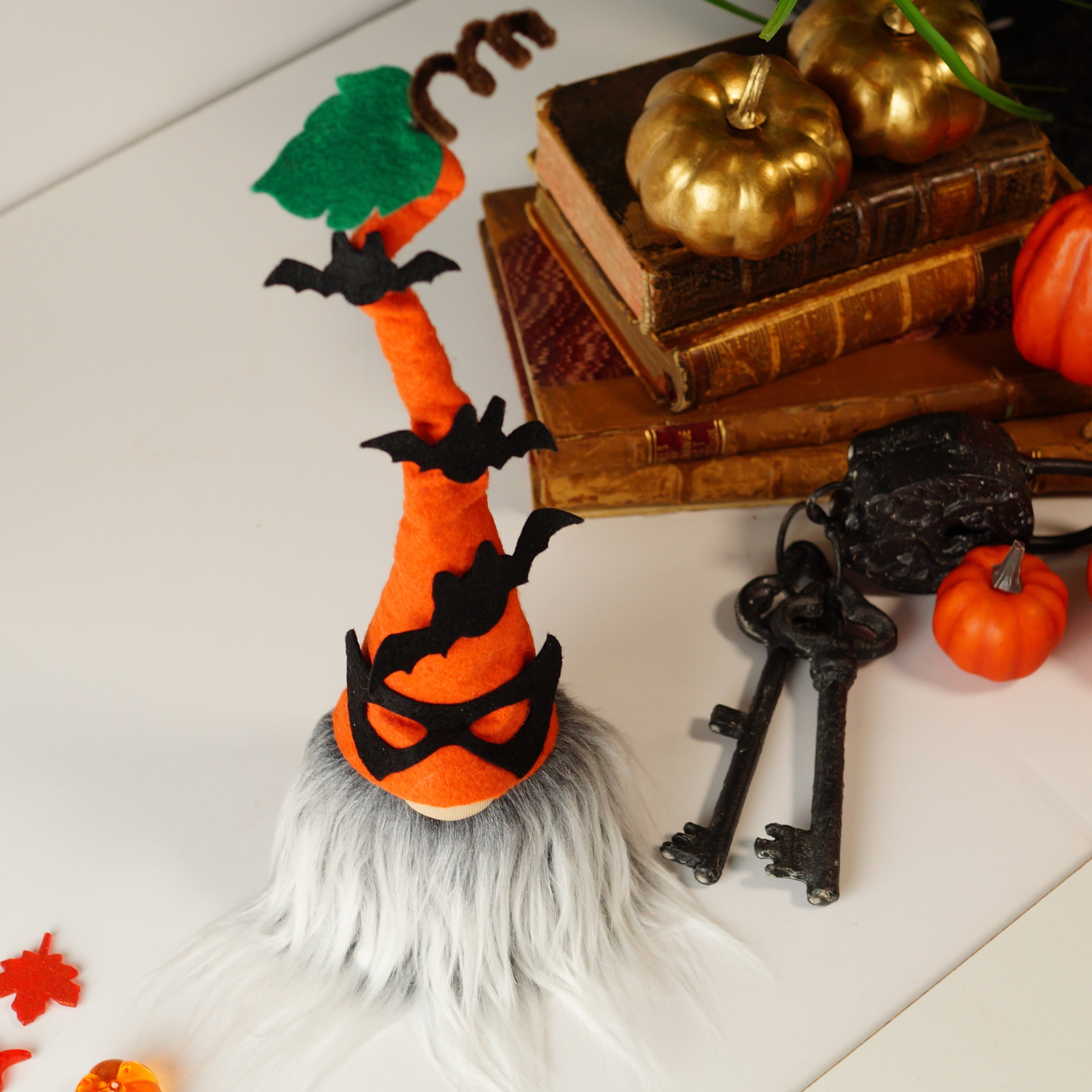 The top view decorative Halloween gnome, dressed in an orange outfit and wearing an orange hat adorned with bats, against a white background with books, pumpkins and a skull.