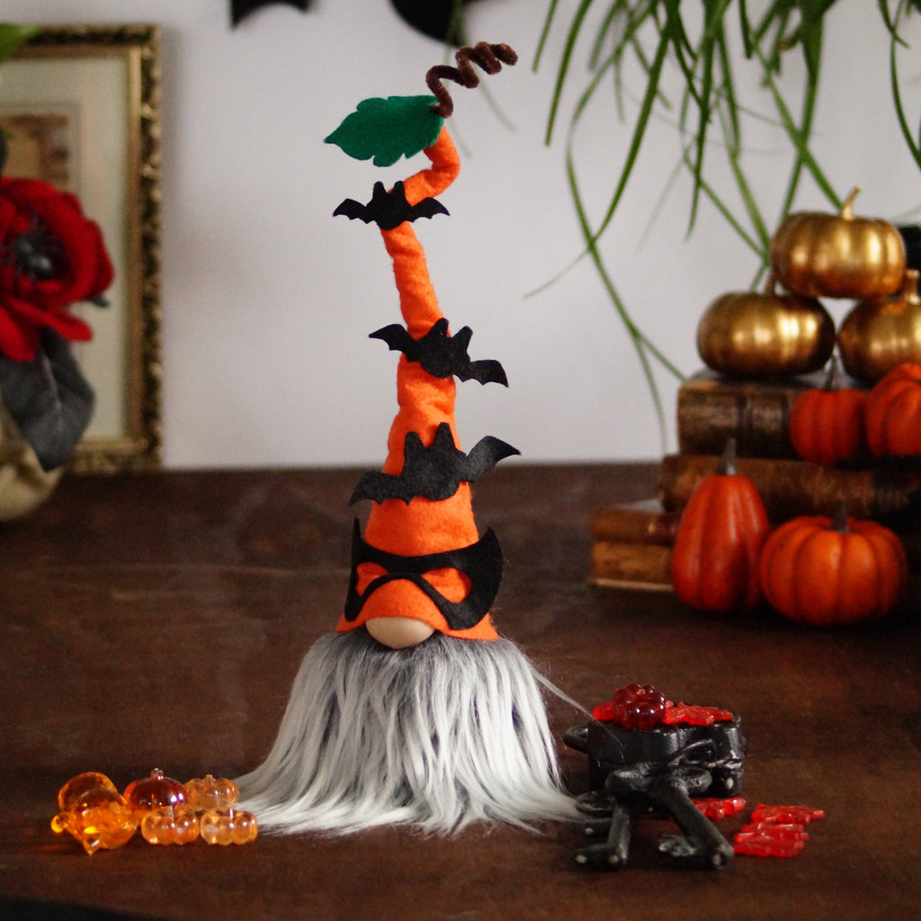 A decorative Halloween gnome, dressed in an orange outfit and wearing an orange hat adorned with bats, against a background with books, pumpkins and a skull.