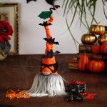 Load image into Gallery viewer, A decorative Halloween gnome, dressed in an orange outfit and wearing an orange hat adorned with bats, against a background with books, pumpkins and a skull.
