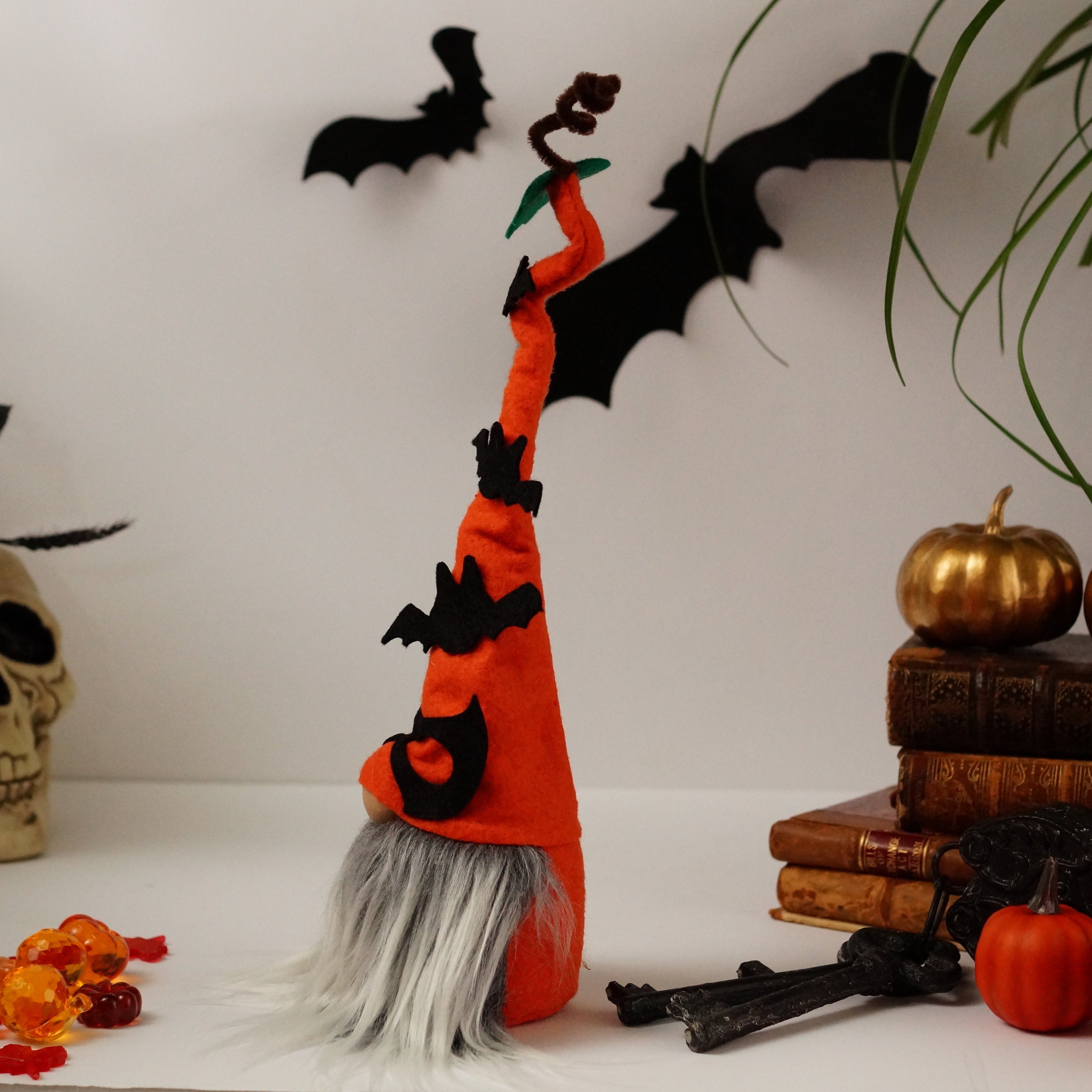 The side view decorative Halloween gnome, dressed in an orange outfit and wearing an orange hat adorned with bats, against a white background with books, pumpkins and a skull.