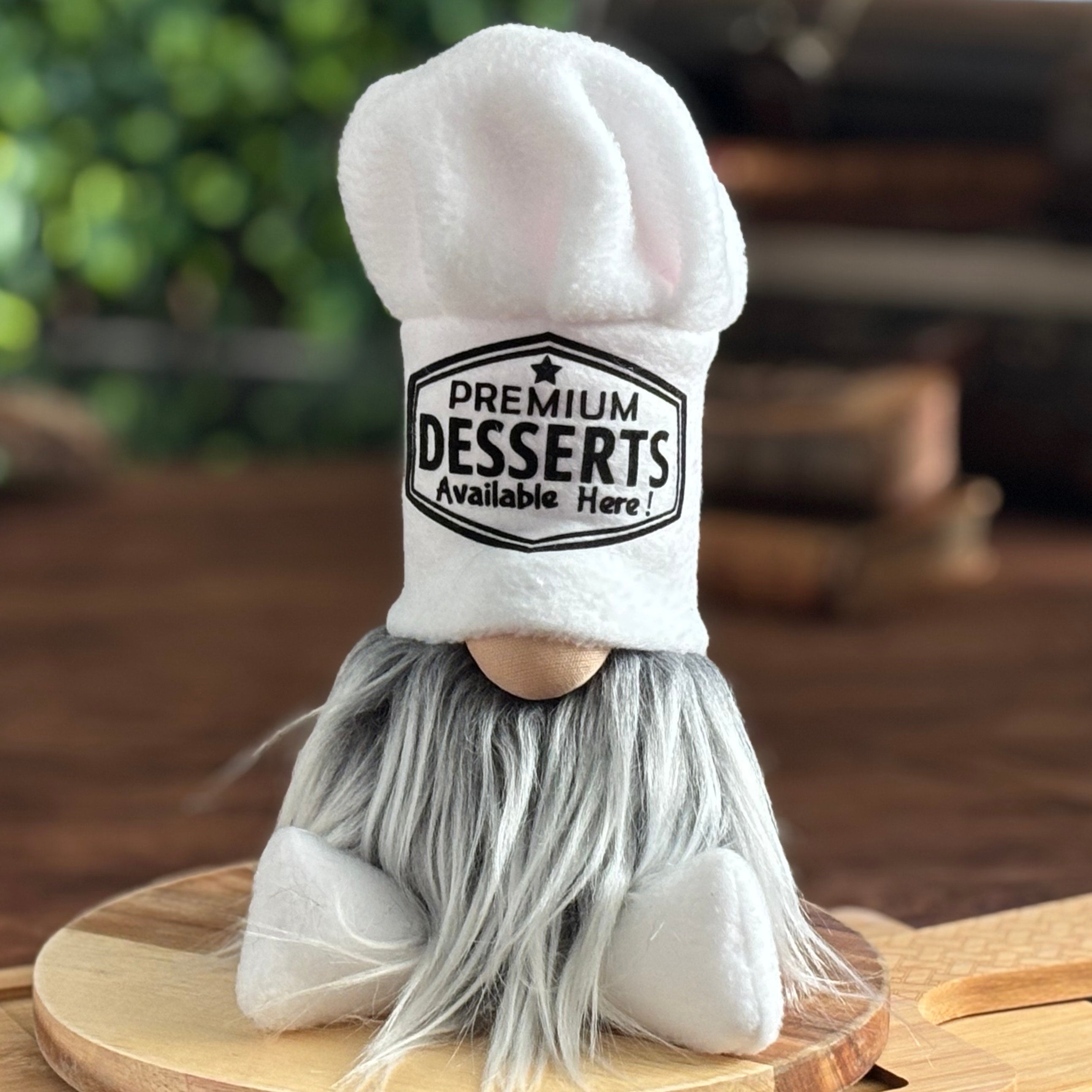 A plush gnome with a long gray beard and a large white chef hat that reads Premium Desserts Available Here sits on a round wooden cutting board. 