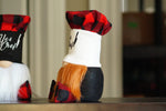 Load image into Gallery viewer, A close-up of a decorative gnome doll with a red and black buffalo plaid hat, a white face band, orange hair, and a black body, standing on a wooden surface.
