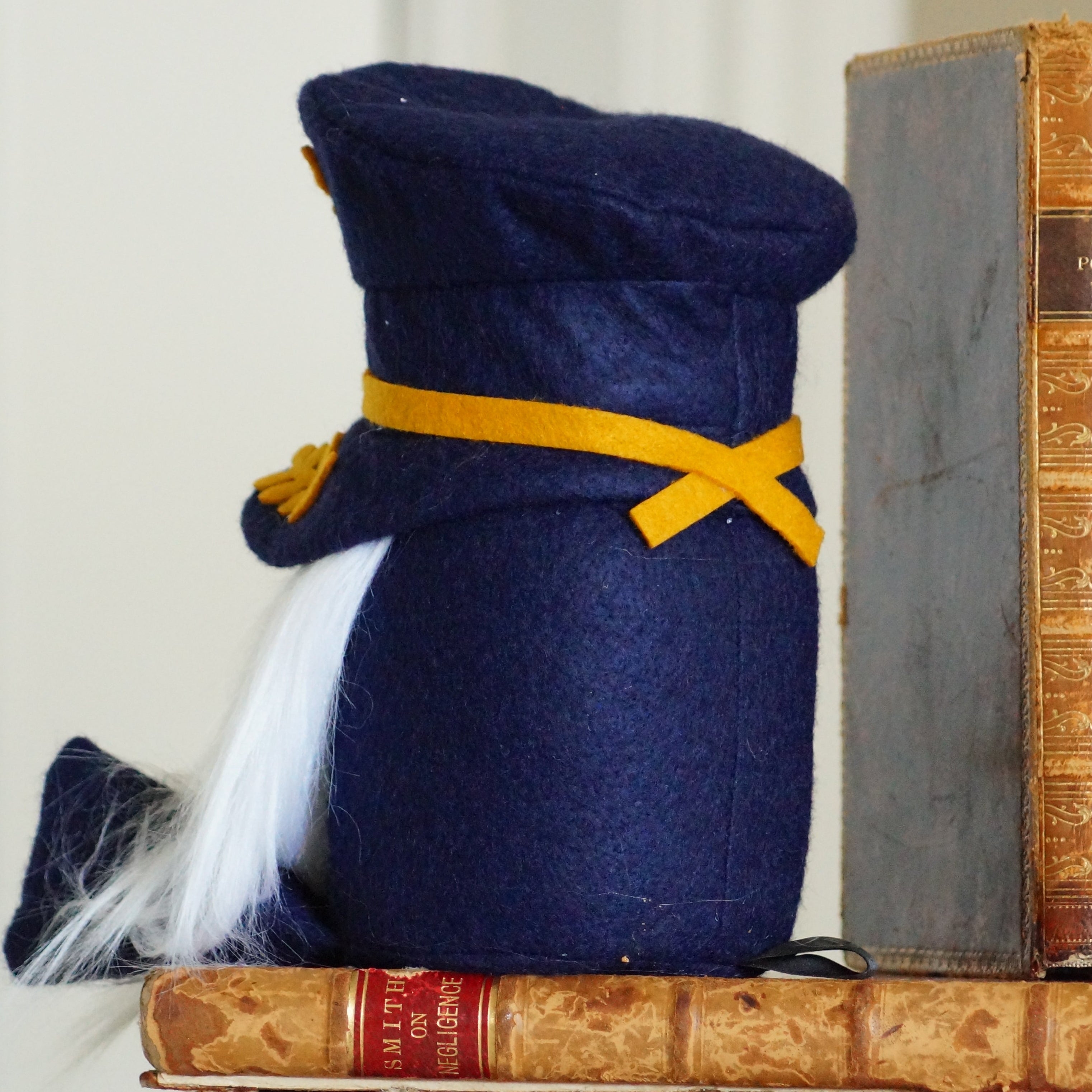 Small plush toy resembling a penguin wearing a navy blue hat with a yellow band, sitting on top of vintage books.