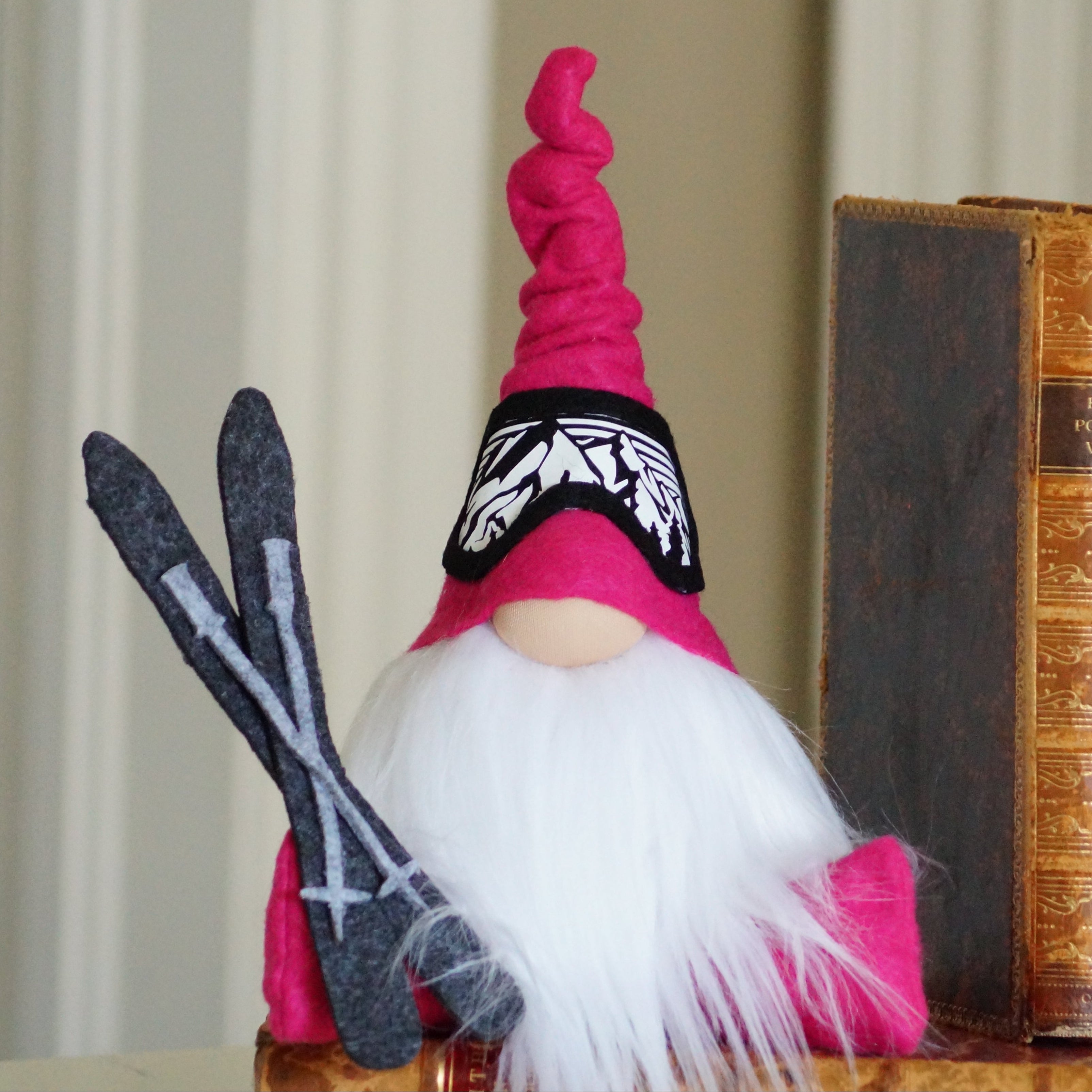 Gnome figurine with pink hat and white beard next to a stack of books on a neutral background with ski goggles and a set of skies