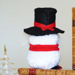 Load image into Gallery viewer, Small snowman figurine with a black top hat and red scarf on a surface with books and a small Christmas tree.
