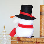 Load image into Gallery viewer, Plush snowman with a top hat and red scarf sitting on books with a Christmas tree in the background.
