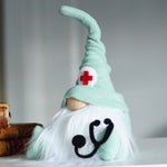 Load image into Gallery viewer, Gnome with a green hat featuring a red cross and a stethoscope symbol, sitting on a surface with books in the background.
