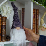 Load image into Gallery viewer, Hand holding a gnome figurine next to books on a decorative shelf with plants and flowers.
