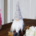 Load image into Gallery viewer, Decorative gnome figure with a white beard and hat on a wooden surface with flowers and other figures in the background.
