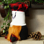 Load image into Gallery viewer, A rustic Christmas gnome doll with a red and black buffalo plaid hat, a white body with black script embroidery, a bright orange beard, and black pants, styled on a shelf with pinecones and greenery.
