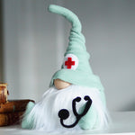 Load image into Gallery viewer, Healthcare worker gnome with a green hat, a red cross, and a stethoscope, sitting near a stack of medical books ideal for Medical white coat ceremony gift

