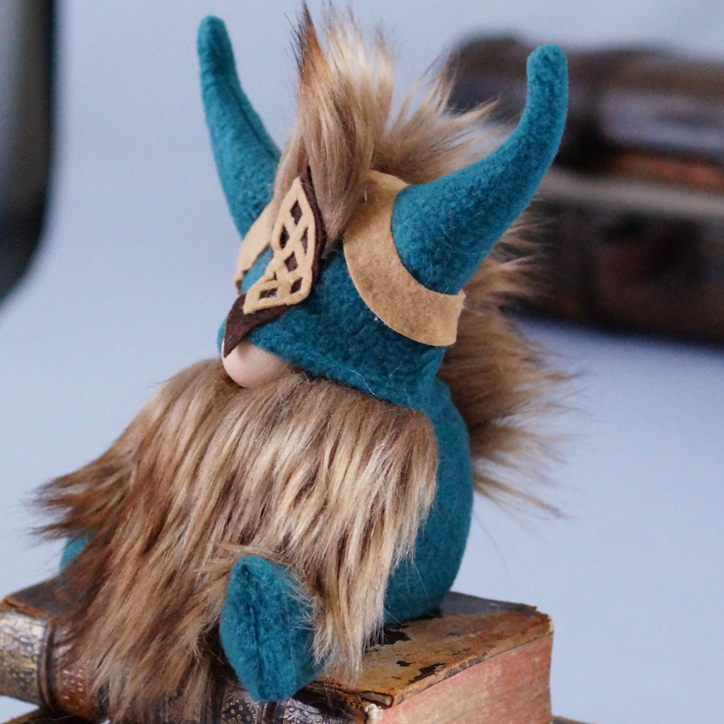 A plush  Viking gnome with a long brown beard and matching mohawk, teal horned helmet, and tan headband sits on a stack of old books, with a vintage suitcase blurred in the background.
