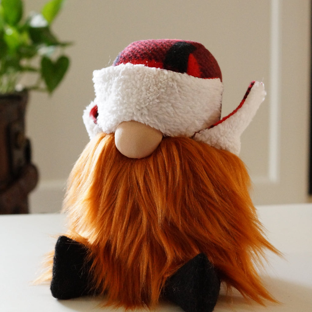 A plush gnome doll with a long ginger beard, a round nose, a red and white buffalo-check lumberjack hat covering its eyes, and black felt boots, sitting against a white background.