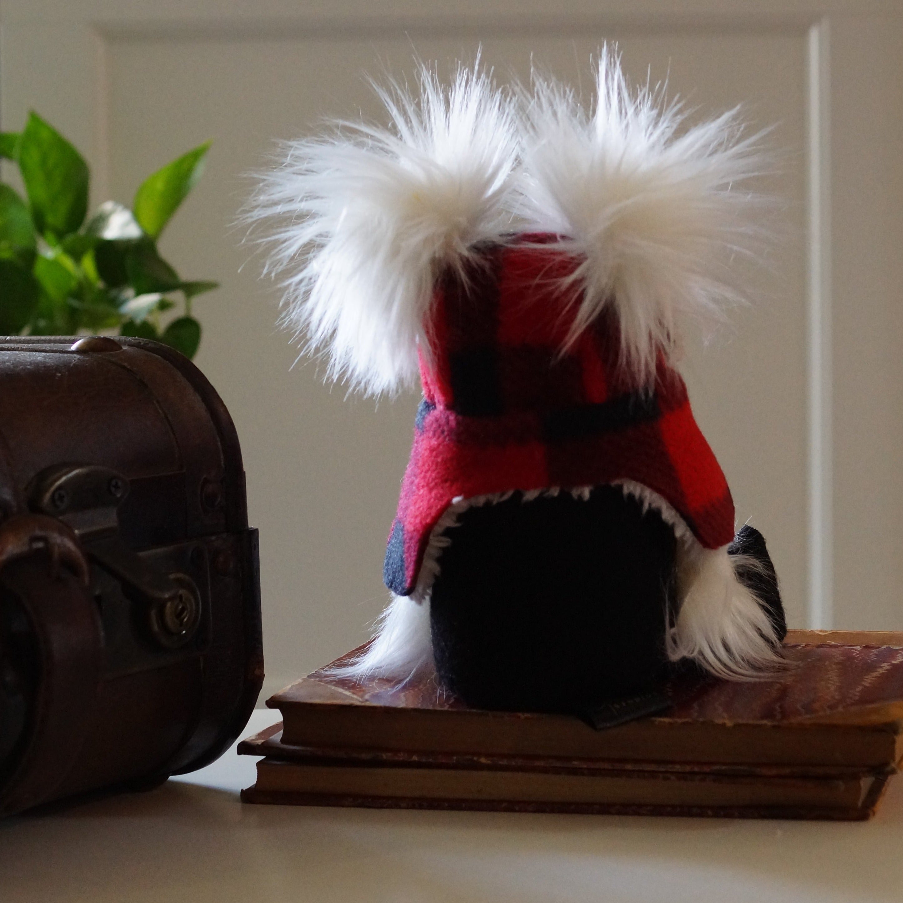A plush Cottage Guardian with white fur and a plaid Lumber Jack hat sits on books by a vintage chest and a green plant.