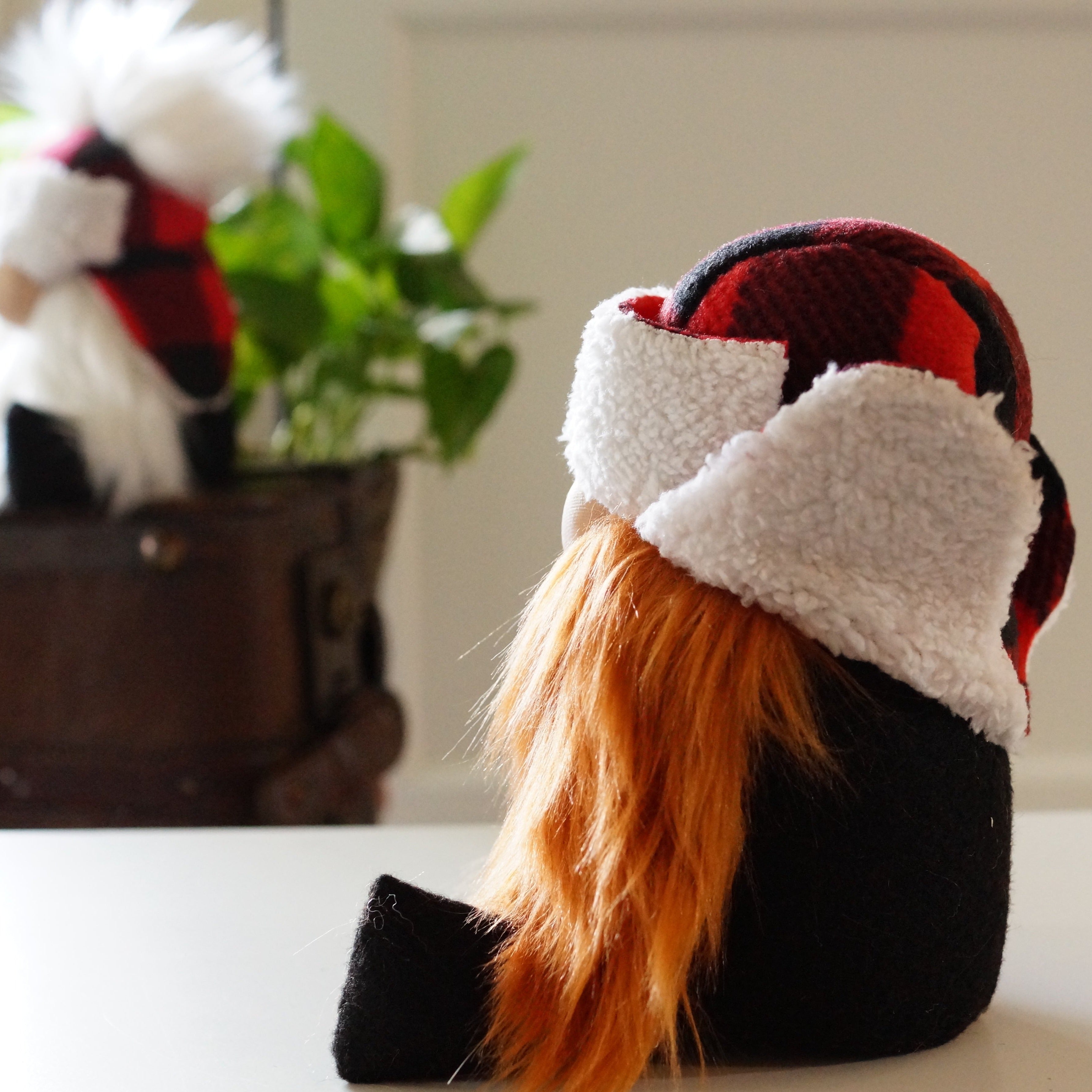 A plush gnome with long orange hair and a red plaid hat stands as a whimsical cottage guardian, with a plant and wooden box nearby.