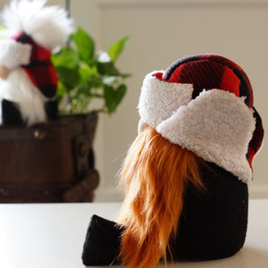 A plush gnome with long orange hair and a red plaid hat stands as a whimsical cottage guardian, with a plant and wooden box nearby.