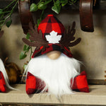 Load image into Gallery viewer, A festive Christmas gnome decoration with a red and black buffalo plaid hat, brown antlers, a long white beard, and red-and-black plaid shoes, displayed on a shelf with a pinecone and holiday decor.
