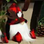 Load image into Gallery viewer, A festive felt gnome decoration wearing a red and black buffalo plaid hat with a white maple leaf, brown felt antlers, and a long white beard, styled on a shelf with a pinecone and greenery.
