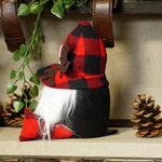 Load image into Gallery viewer, A small decorative gnome figurine with a red and black buffalo plaid hat, white beard, and red shoes, displayed on a shelf with pinecones and greenery.
