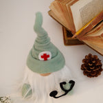 Load image into Gallery viewer, Doctor Gnome figurine with a green hat and red cross symbol next to an open book and pine cone on a white surface. Ideal gift for the Doctor reception 
