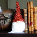 Load image into Gallery viewer, Orange Gnome with white faux fur sitting on a shelf next to vintage books and a suitcase.
