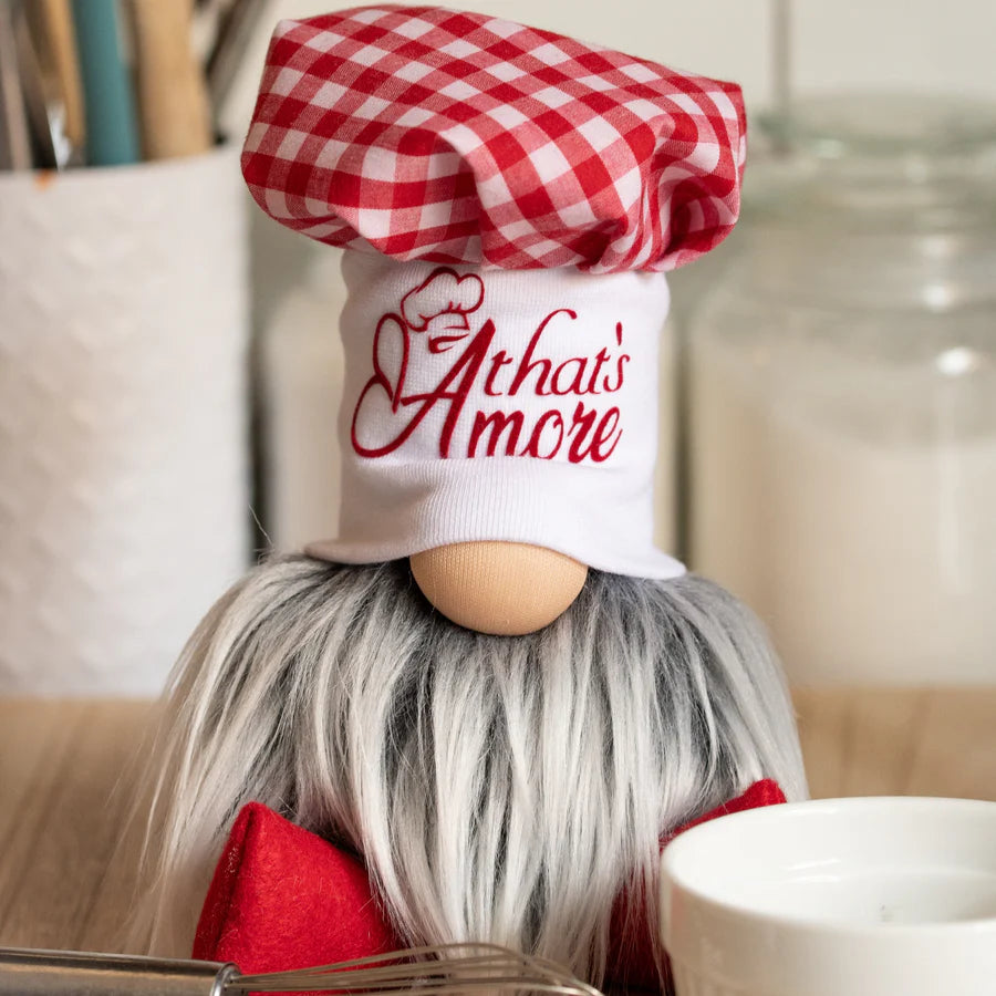 A handmade kitchen guardian, Papa Giuseppe, with a white beard and red checked hat sits on the counter near utensils and a candle.