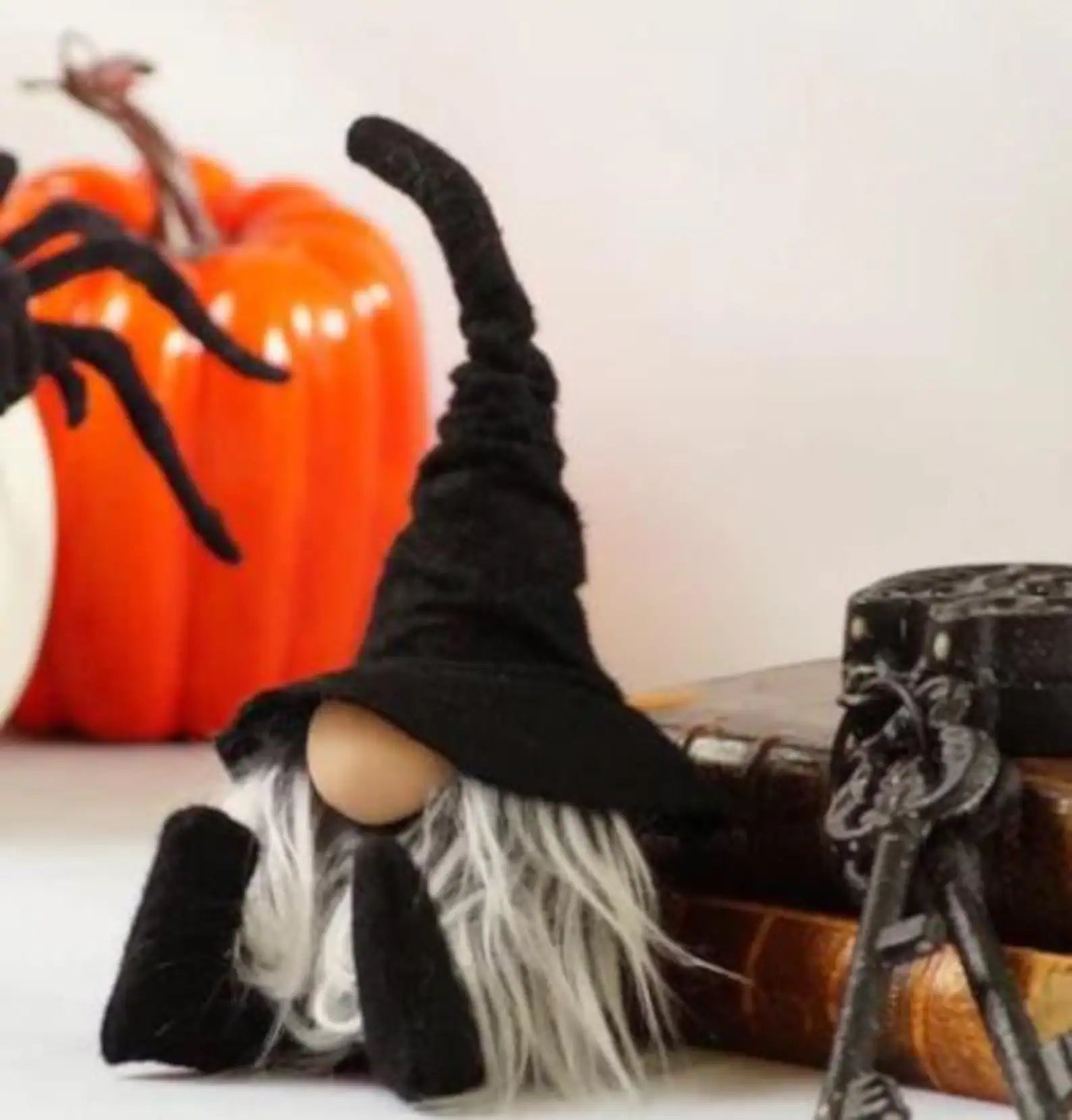 A gnome with a tall black hat and gray beard sits beside two old books with a skeleton key and lock on top. In the background are an orange pumpkin, a white pumpkin, a black spider, and a black-and-red artificial flower.