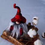 Load image into Gallery viewer, A handmade library guardian gnome with a red plaid hat and gray beard sits on vintage books beside cotton branches and pinecones.
