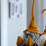 Load image into Gallery viewer, A handmade plush gnome with a long white beard and tall yellow hat sits sideways indoors, perfect as a charming home office or library guardian.
