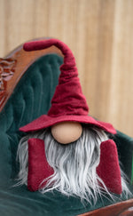 Load image into Gallery viewer, A plush gnome with a tall red hat and gray beard sits on a green velvet chair, perfect as a handmade library guardian for your home office or study.
