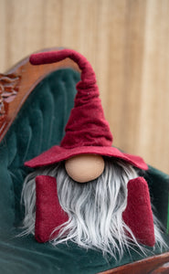 A plush gnome with a tall red hat and gray beard sits on a green velvet chair, perfect as a handmade library guardian for your home office or study.