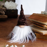 Load image into Gallery viewer, Small gnome figurine with a brown hat and white beard on a wooden surface with books and flowers.
