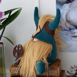 Load image into Gallery viewer, Plush toy with blue hair and horns on a wooden block next to a plant and sculpture.
