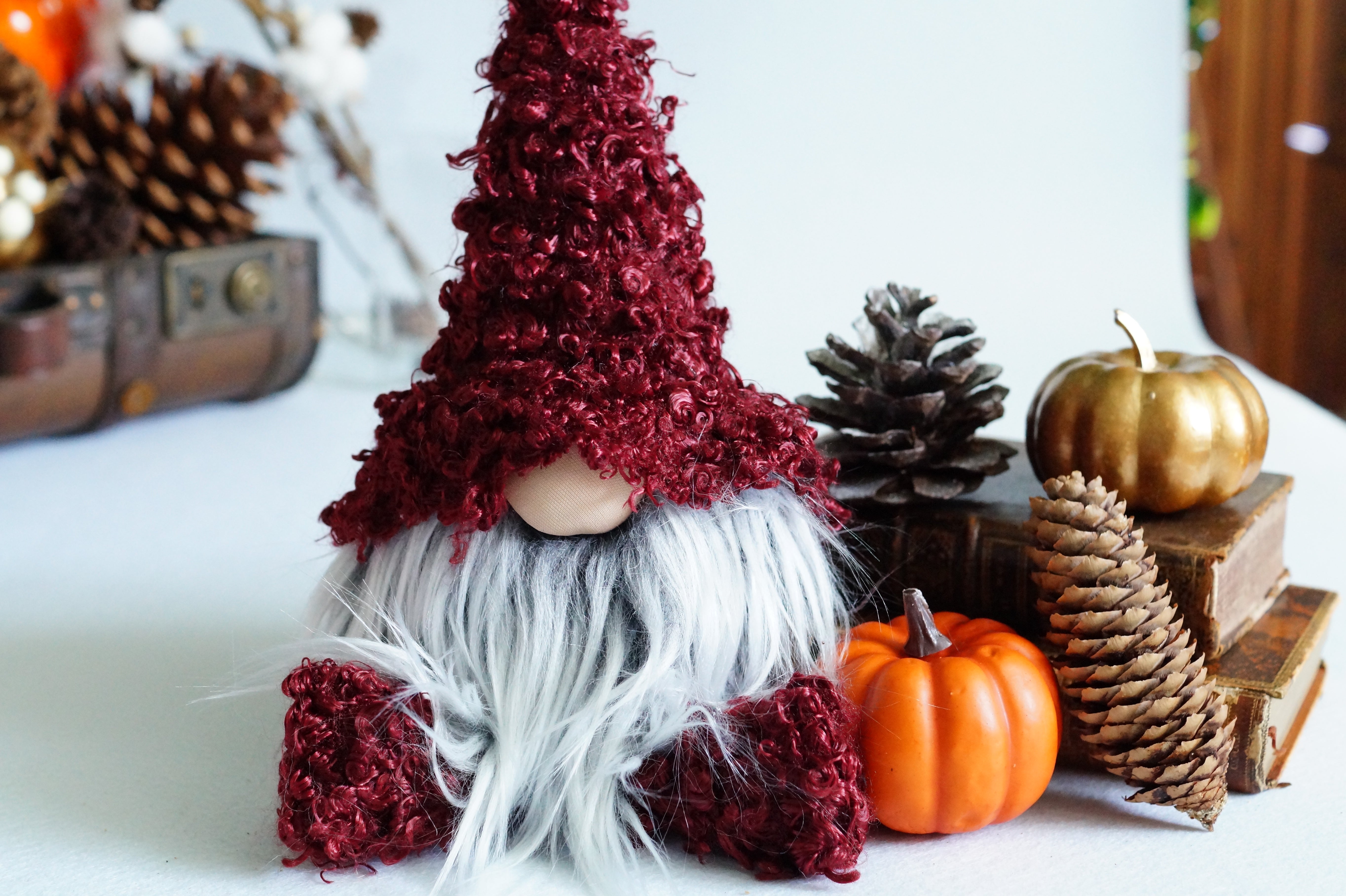 Gnome with a red hat and white beard surrounded by pumpkins and pinecones on a white surface.