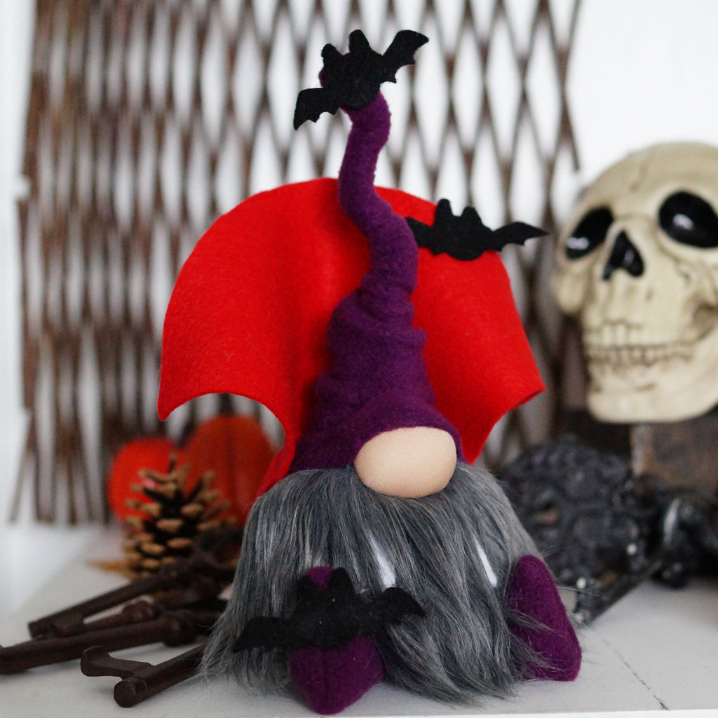 A purple vampire gnome with a bendable hat, sitting beside a decorative skull and pine cones, designed for Halloween decor.