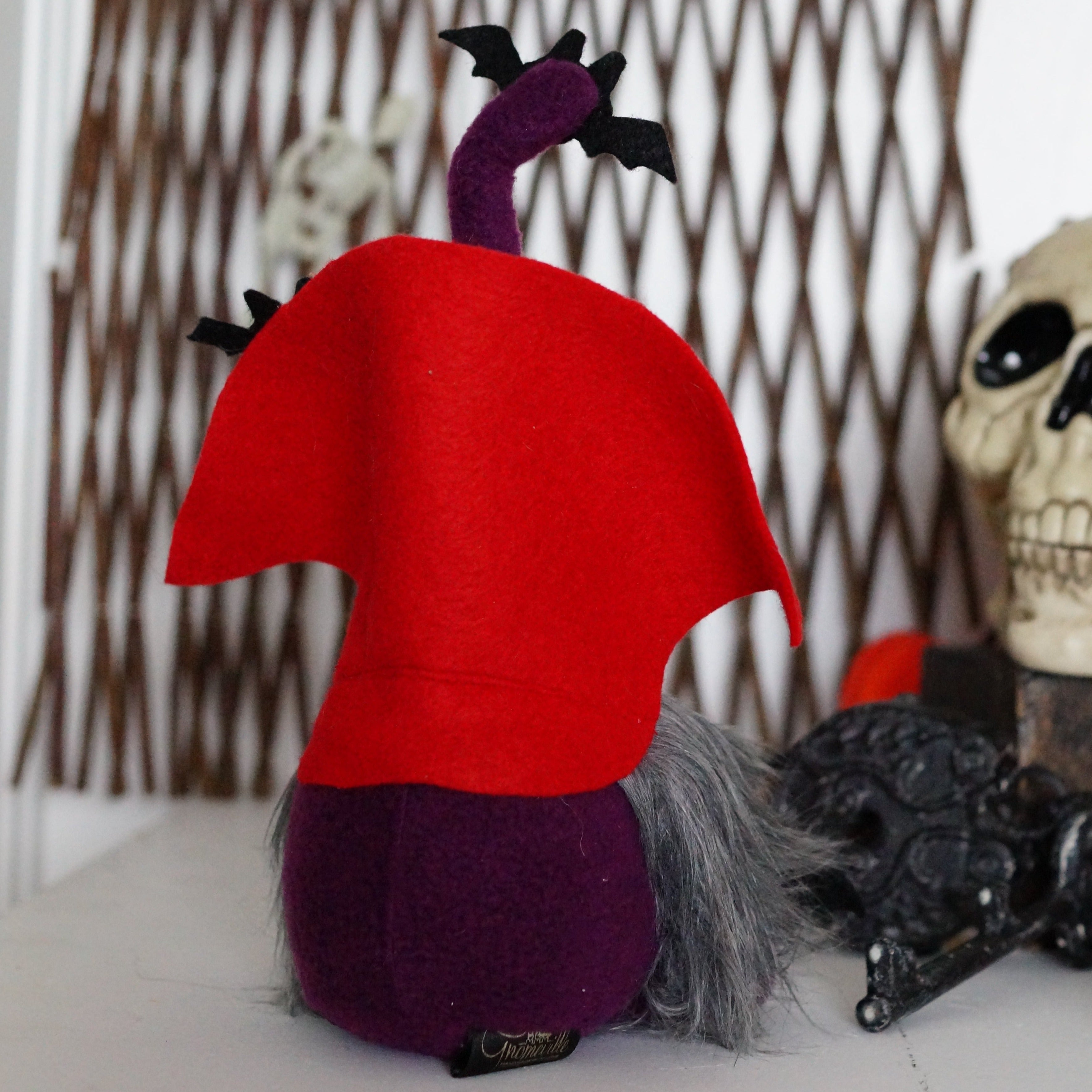 The back view of a purple vampire gnome with a bendable hat, sitting beside a decorative skull and pine cones, designed for Halloween decor.
