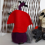 Load image into Gallery viewer, The back view of a purple vampire gnome with a bendable hat, sitting beside a decorative skull and pine cones, designed for Halloween decor.

