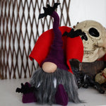Load image into Gallery viewer, A purple vampire gnome with a bendable hat, sitting beside a decorative skull and pine cones, designed for Halloween decor.
