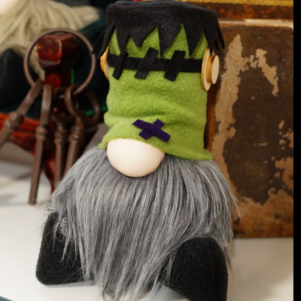 A handmade gnome with a green head resembling Frankenstein's monster, with a black patch over one eye and a wooden screws on the hat. The gnome has a gray beard and is seated against a decorative background.