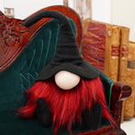 Load image into Gallery viewer, handmade Halloween collectible fabric gnome wearing black fleece wizard hat and red beard for gothic tiered tray vibes sitting on a couch with books antique  in the back ground
