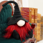 Load image into Gallery viewer, Handmade collectible fabric gnome wearing a black fleece wizard hat and red beard sitting on a couch next to antique books perfect for Halloween decor
