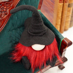 Load image into Gallery viewer, A handmade library guardian gnome doll with red hair and a black hat sits on vintage books beside a green velvet chair—perfect for your study.
