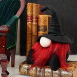 Load image into Gallery viewer, A handmade library guardian gnome doll with red hair and a black hat sits on vintage books beside a green velvet chair—perfect for your study.
